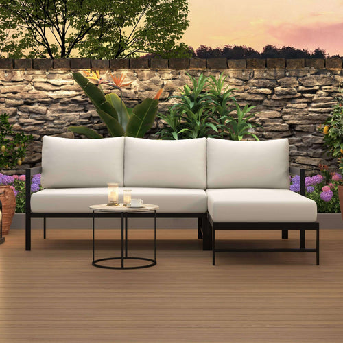 A 3-seater outdoor sectional sofa with white cushions and a black metal frame, shown with a matching chaise lounge and a small round side table in a staged patio setting with a stone wall, plants, and a wooden floor.